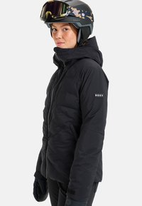 Black puffer jacket with a hood, featuring quilted texture, adjustable cuffs, and a Roxy logo on the sleeve. Worn with a black ski helmet and goggles.