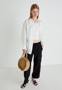Witte button-down shirt over een witte geribbelde crop top, gecombineerd met zwarte broek. Houdt een ronde gevlochten tas vast; zwarte sandaaltjes met hakken en banden.