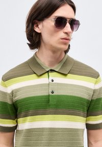 Νέος άνδρας με μεσαίου μήκους καστανά μαλλιά που φοράει σκούρα γυαλιά ηλίου και πράσινο πλεκτό πόλο με ρίγες, κοιτάζοντας προς τα δεξιά.