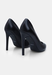 Fekete bőr magassarkú cipők hegyes orral és vékony stiletto sarkakkal. Finom textúrával és egy kis logóval a hátsó részen.