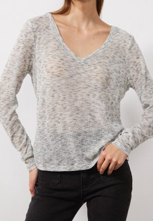 Frau trägt einen hellgrauen, transparenten Pullover mit V-Ausschnitt, langen Ärmeln und einem dezenten Tiermuster sowie schwarze Hosen.