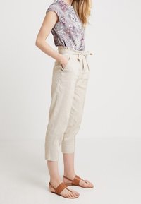Blouse à motifs gris clair à manches courtes, associée à un pantalon en lin beige raccourci noué à la taille, et des sandales plates marron.