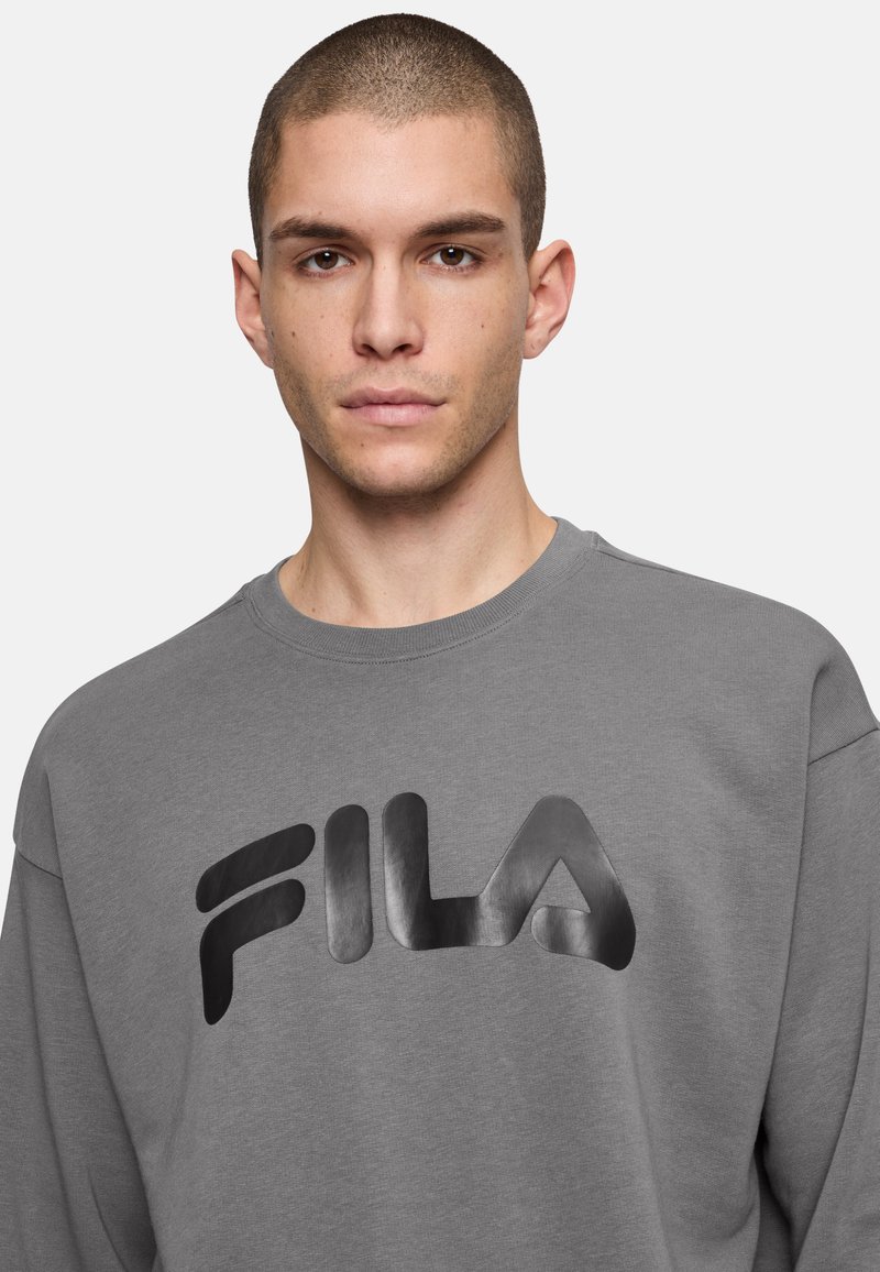 Felpa grigia con collo rotondo, che presenta un grande logo nero FILA sul davanti. Tessuto liscio e vestibilità rilassata.