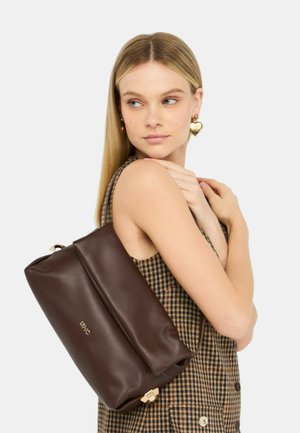 Sac en cuir marron au fini lisse, avec un haut enroulé et des accessoires en or. Porté comme un sac à dos. Robe à carreaux en arrière-plan.