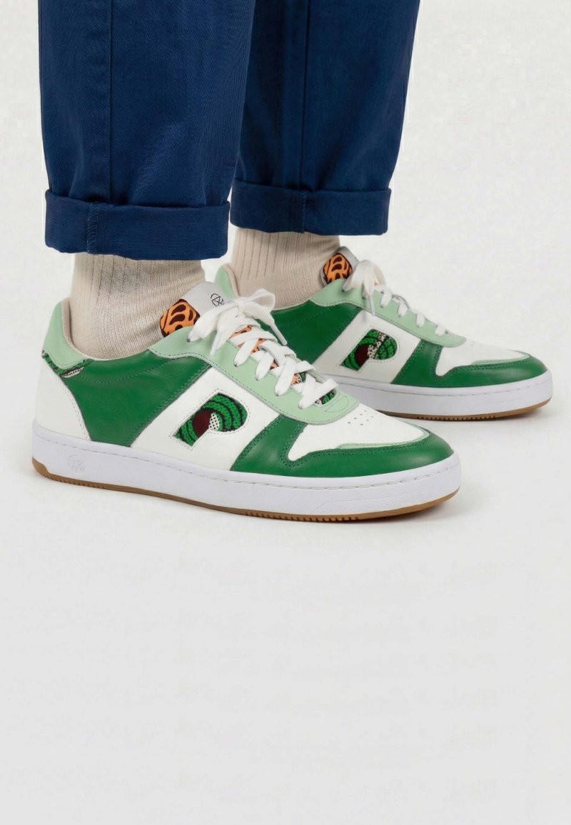 Personne portant des baskets basses vertes et blanches avec un logo à motifs, des chaussettes beiges et un pantalon bleu retroussé, debout sur un fond clair.