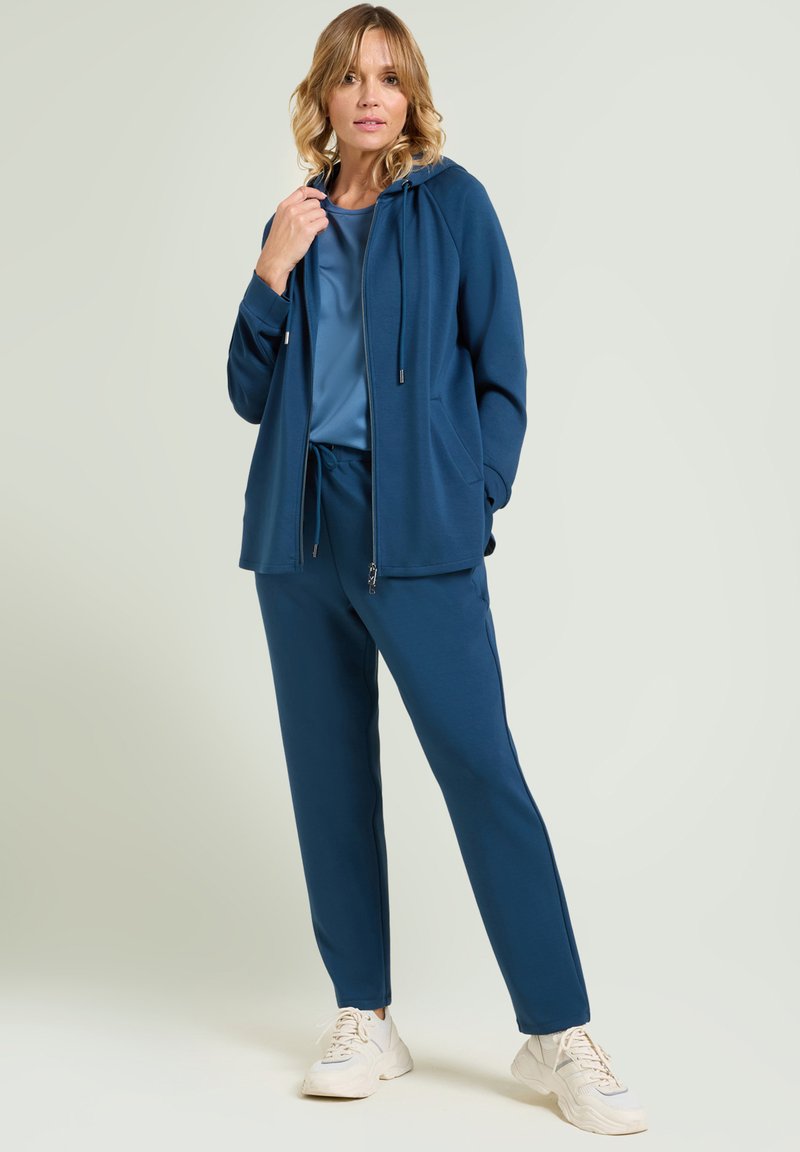 Felpa blu navy con zip e cordino, abbinata a pantaloni jogger coordinati. Texture liscia, vestibilità rilassata. Abbinata a sneaker bianche.