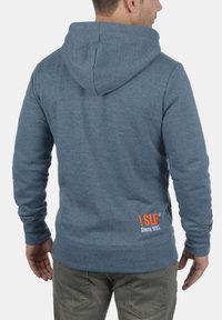 Sudadera con capucha azul hecha de tela suave, con un logo en naranja en la parte posterior, costuras visibles y un corte relajado.