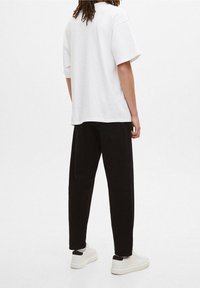 Biała, oversize koszulka z krótkimi rękawami, zestawiona z czarnymi, zwężanymi spodniami i białymi trampkami, na jednolitym tle.