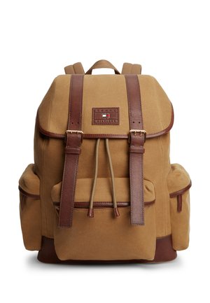 Sac à dos en toile couleur tan avec des bretelles en cuir marron, poches latérales, poche avant à cordon et logo Tommy Hilfiger sur le rabat.