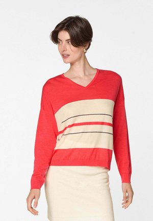 Vrouw die een rood- met crèmegekleurde trui met V-hals en horizontale strepen draagt, gecombineerd met een crèmekleurige rok, poserend tegen een effen achtergrond.