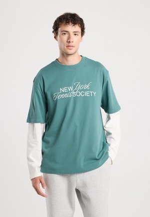 Korte mouw teal t-shirt met witte tekst "New York Tennis Society." Lange mouw witte shirt zichtbaar eronder. Grijze joggingsbroek.