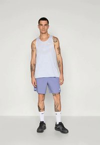 Jonge man met tatoeages die een lichtgrijze mouwloze Nike-top, paarse Nike-shorts, witte Nike-sokken en zwarte sneakers draagt, staande tegen een effen achtergrond.