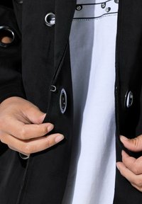 Chaqueta de ante negra con grandes acentos de ojales plateados, con un primer plano de manos ajustando los cierres. Camisa blanca debajo.