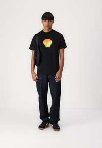Zwarte katoenen T-shirt met een hexagonaal grafisch ontwerp in oranje en groen, gecombineerd met donkerblauwe jeans en zwarte sneakers. Een zwarte schoudertas wordt gedragen.