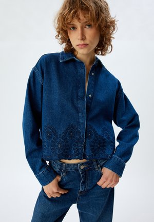 Blue denim cropped shirt with a buttoned collar, featuring intricate dark blue embroidery along the hem and a relaxed fit.