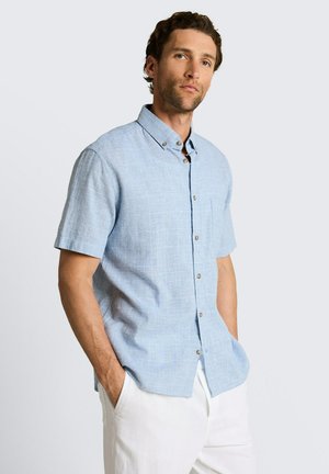 Homme portant une chemise bleu clair à manches courtes avec boutons et poche avant, et un pantalon blanc, debout les mains dans les poches devant un fond uni.