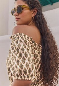 Off-shoulder topp med ett blommönster i guld och svart på en ljusrosa tyg; modell som bär gröna kattröda solglasögon och hoops. 
