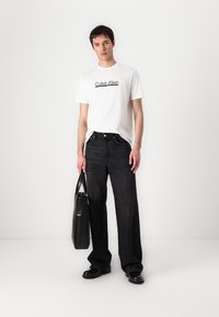Jeans anchos negros, camiseta blanca con logo negro de "Calvin Klein", zapatos negros y una bolsa tote negra. Fondo neutro.