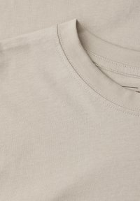 Gros plan sur un t-shirt beige à col rond montrant le col et la partie supérieure de la poitrine avec des détails de couture visibles.