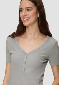 Lichtgrijze, geribbelde top met korte mouwen, een v-hals en decoratieve knopen aan de voorkant. Zachte textuur, slim fit ontwerp en subtiele accenten bij de halslijn.