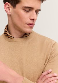 Maglione beige a maglia con collo rotondo, caratterizzato da un colletto a righe in contrasto. Dettaglio logo in rilievo sul petto, texture liscia.