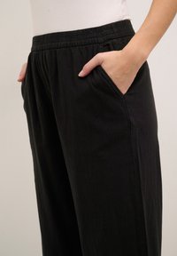Black, loose-fitting pants made of soft fabric, featuring a wide elastic waistband and side pockets. Smooth texture with no visible patterns.