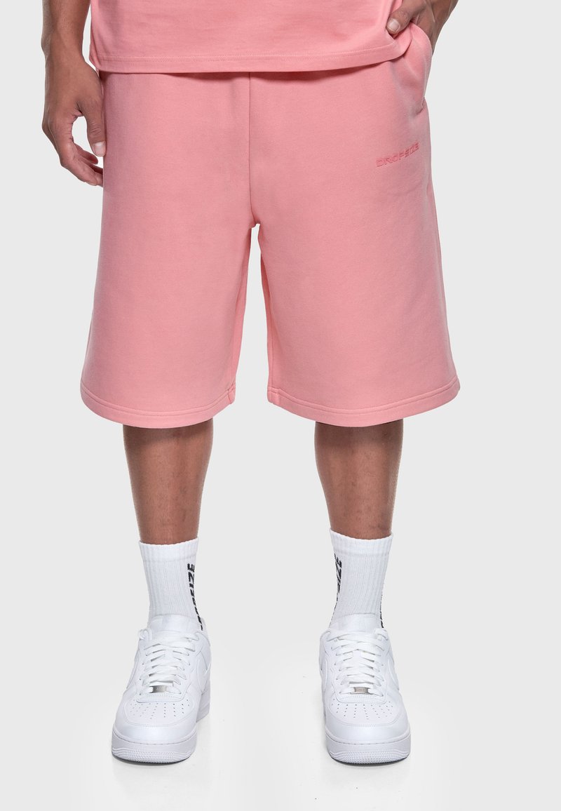 Homme portant un short rose mi-long, des chaussettes blanches montantes avec un texte noir et des baskets blanches, debout sur un fond clair.