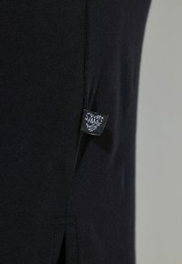 Černá tkanina s hladkým povrchem, s bočním švem a malou cedulkou s tištěným logem. Jednoduchá a minimalistická estetika.