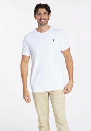T-shirt en coton blanc à manches courtes, avec un petit logo noir sur la poitrine gauche. Porté avec un pantalon beige clair.