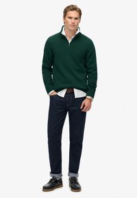Suéter verde de punto con cuello de media cremallera, combinado con jeans azul oscuro y zapatos negros con cordones, con puños enrollados y un cinturón marrón.