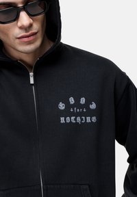 Felpa nera con zip e grafica grigia che recita "GOOD for NOTHING." Presenta un cappuccio, tasche frontali e una texture morbida.