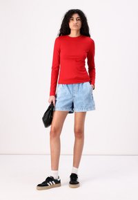 Top roșu cu mânecă lungă combinat cu pantaloni scurți din denim deschis.Ținuta include pantofi sport negri cu accente albe și o geantă neagră.