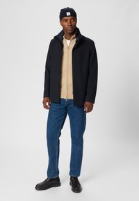 Marinblandad ullrock med knäppning, beige zip-up tröja, blå denimjeans och svarta läderskor. Svart stickad mössa på huvudet.