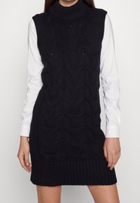 Abito in maglia nera con orlo a coste, dotato di un collo alto e motivo a trecce testurizzato. Camicia bianca a maniche lunghe sotto.