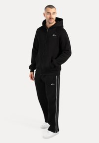 Sort zip-up hættejakke og matchende sweatpants lavet af blødt stof. Hættejakken har et fremtrædende logo, og bukserne har sidelange striber og lommer. Hvite sneakers.