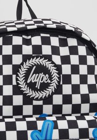Czarny i biały plecak w kratkę z haftowanym okrągłym logo z napisem "hype." oraz wzorem wawrzynu. Wyraźne niebieskie akcenty graficzne.