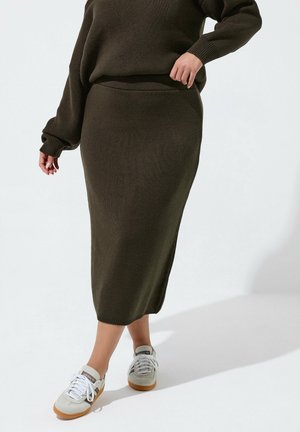 Kvinde iført mørke olivengrøn strikket midi-nederdel og matchende sweater, stående i hvide sneakers på hvid baggrund.
