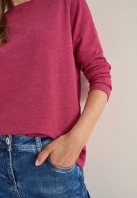 Czerwona w paski bluzka z długim rękawem o luźnym kroju, zestawiona z niebieskimi dżinsami. Bluzka ma okrągły dekolt i podwinięte rękawy.