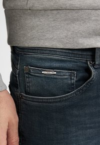 Primer plano de un bolsillo de unos vaqueros de mezclilla azul oscuro con una pequeña etiqueta metálica "Petrol Ind" y una mano descansando junto a la cadera, llevando un suéter gris.