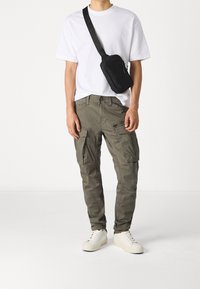 Olivgröna cargobyxor med flera fickor, kombinerade med en enkel vit T-shirt och en svart axelväska. Strukturerad tyg, lös passform.