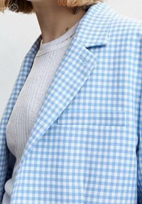 Blazer a quadre blu chiaro con rever, taschino sul petto e una texture sottile, indossato sopra un top grigio a coste.