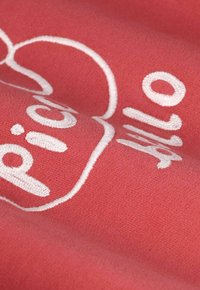 Un textile corail présentant un texte brodé blanc dans une police ludique, avec une texture lisse et un léger éclat. Le tissu semble doux et léger.