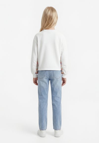Παιδί με μακριά ξανθά μαλλιά, που φοράει ένα λευκό πουλόβερ με διακοσμητικές πλαϊνές λωρίδες, ανοιχτό μπλε τζιν και λευκά αθλητικά παπούτσια, γυρισμένο προς τα πίσω.