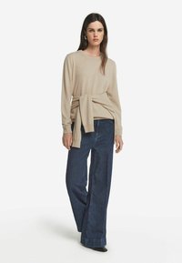 Beżowy sweter z długim rękawem i wiązaniem w talii, zestawiony z luźnymi ciemnymi dżinsami i wsobnymi czarnymi butami, na białym tle.