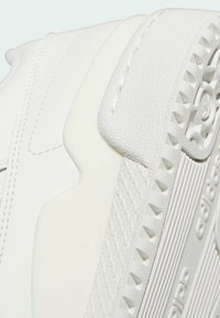Close-up da sola e lateral de um tênis Adidas branco, mostrando o grip texturizado, costura e o logotipo em relevo da Adidas no entressola.