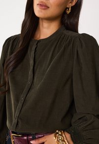 Blusa in velluto a coste verde scuro con collo rotondo, chiusura a bottoni e maniche lunghe leggermente a sbuffo con dettagli in pizzo. Abbinata a una cintura.