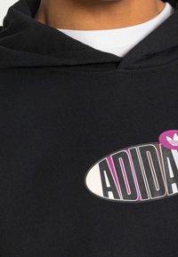 Svart hoodie med en stor oval logotyp med "ADIDAS"-text i olika färger, inklusive rosa och lila detaljer, på ett mjukt tyg.