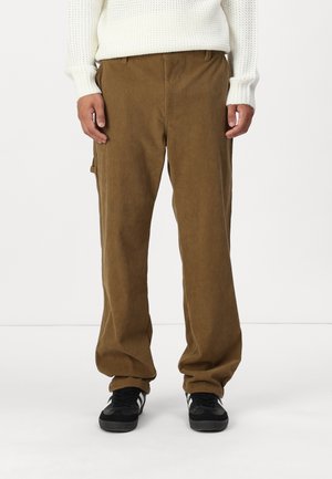 Braune Cordhosen mit weitem Bein, die eine vertikale Rippstruktur und eine Seitenasche aufweisen. Kombiniert mit schwarzen Sneakers.
