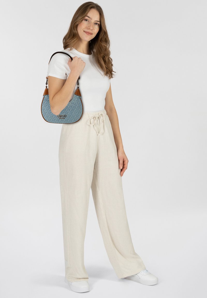 Borsa azzurro chiaro con tessuto decorato e dettagli in pelle marrone, indossata con una maglia bianca aderente e pantaloni beige a gamba larga, su uno sfondo neutro.