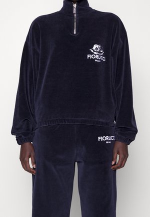 Granatowy welurowy dres z półrozpinanym pulowerem i dopasowanymi spodniami, oba z białymi logo Fiorucci Milan na klatce piersiowej i udzie.